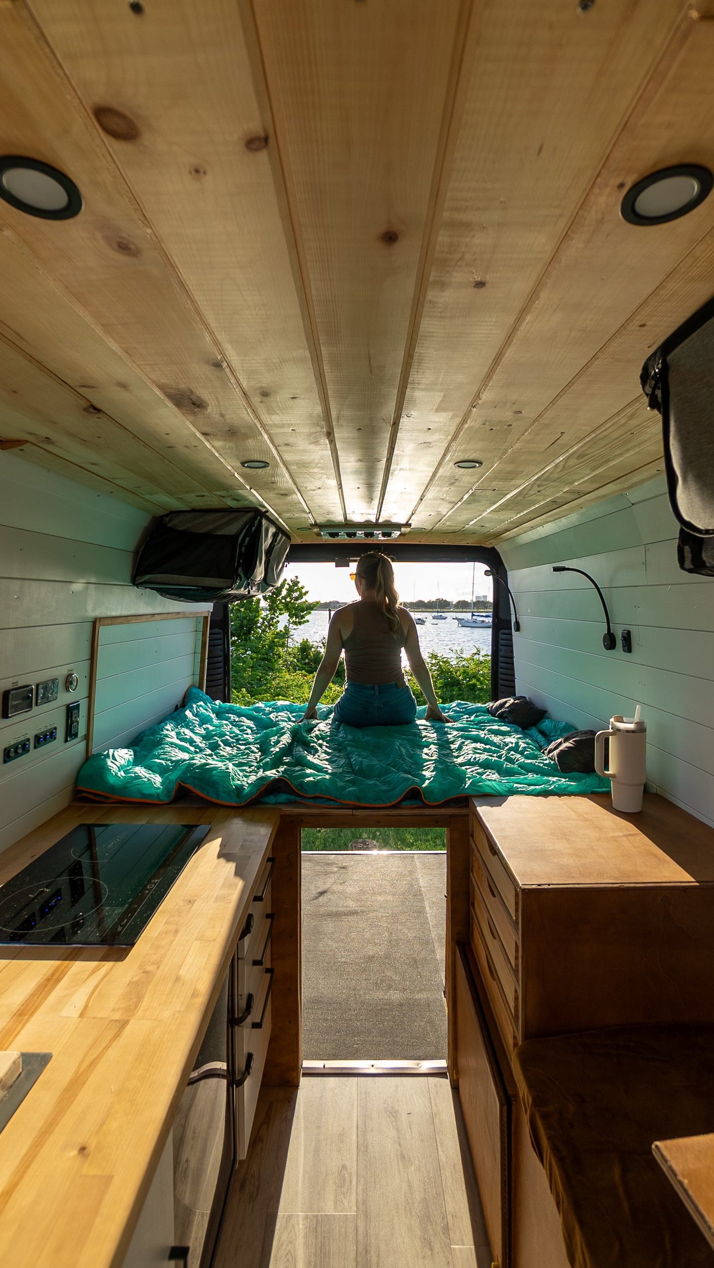 Cozy rv rental van life setup. Woman on bed overlooks scenic water with boats. Features wooden ceiling, kitchen, and custom storage.