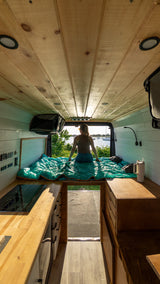 Cozy rv rental van life setup. Woman on bed overlooks scenic water with boats. Features wooden ceiling, kitchen, and custom storage.