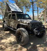 Custom Jeep Gladiator with a pop-top camper, large off-road tires, parked in a sunny forest. Perfect RV rental for adventures!