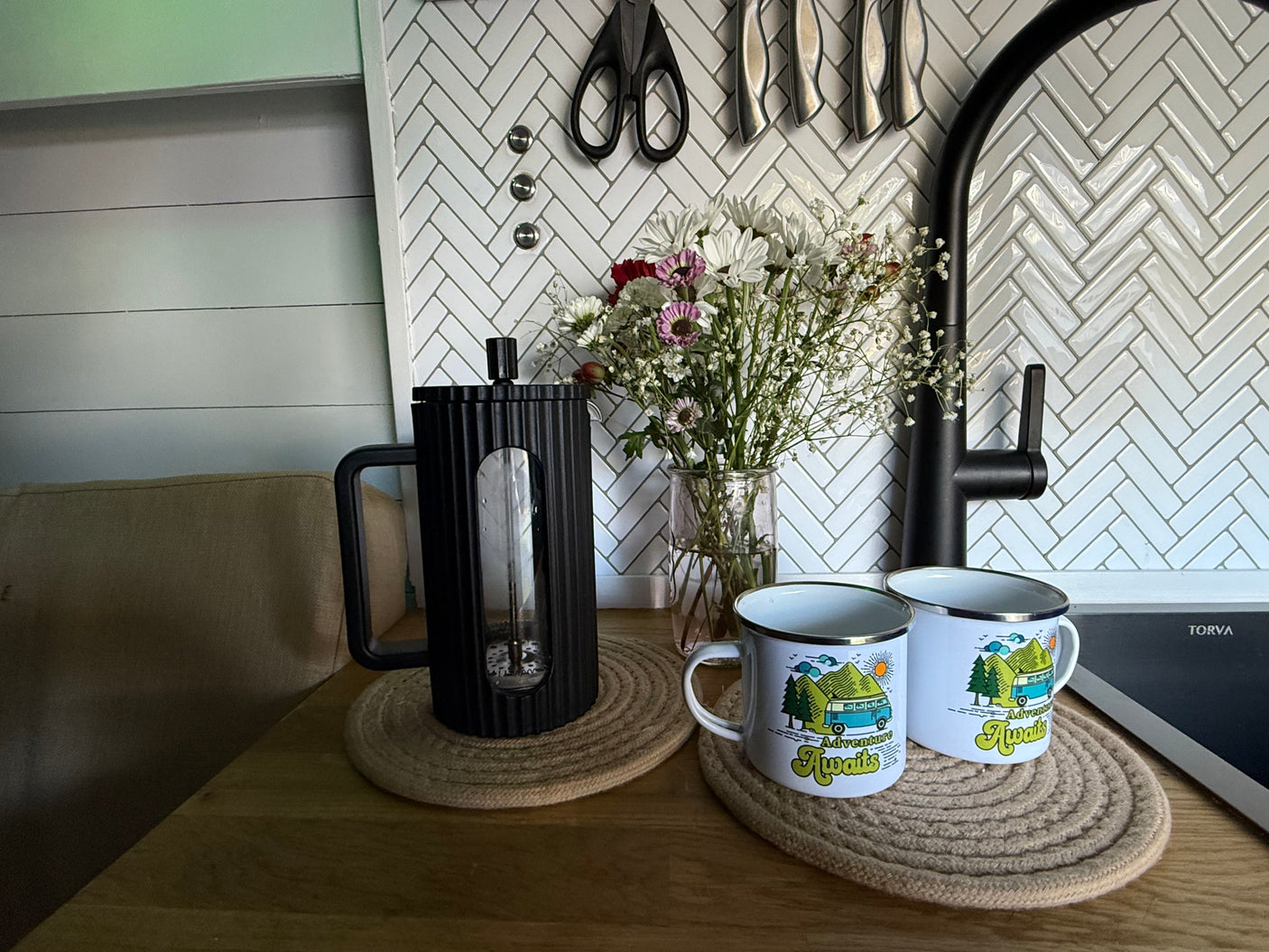An rv rental kitchen view: fresh flowers, a French press, and "Adventure Awaits" mugs, all set on a cozy wooden counter.