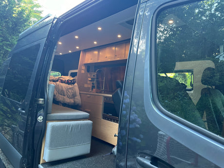 Modern RV rental camper van interior: open door shows kitchen, seating/bed area, and ceiling lights.