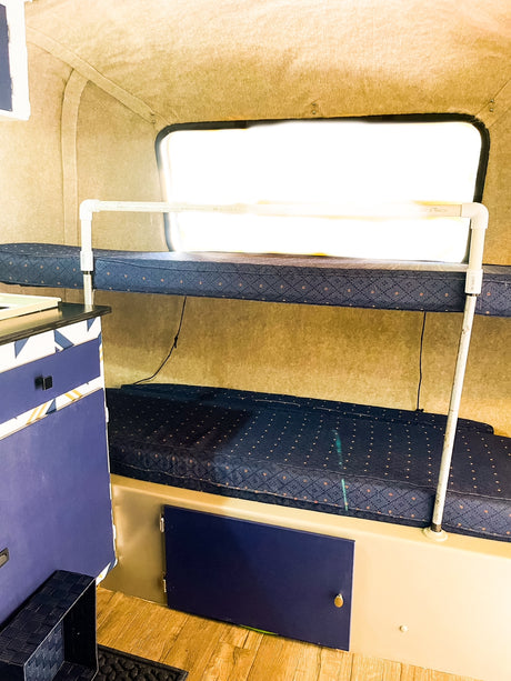 Compact RV rental interior featuring two bunk beds with blue mattresses, white PVC safety rail, and lower storage.