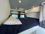 RV rental interior: A cozy bedroom with a dark blue bed, overhead storage, ambient lighting, and rear windows.