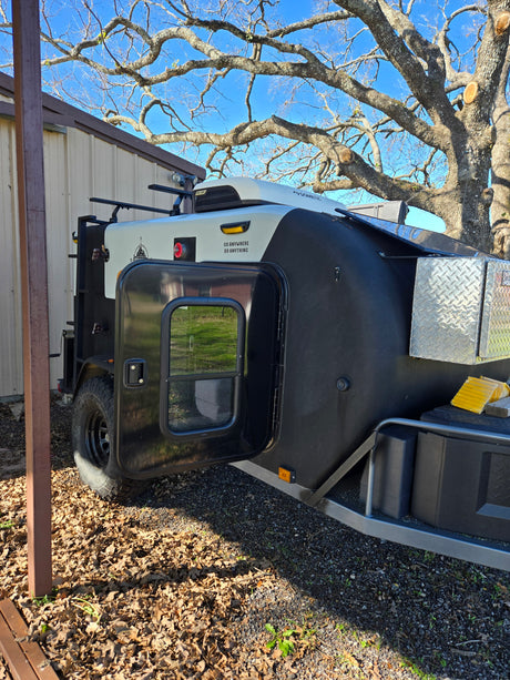 Black & white off-road teardrop camper trailer, ideal for RV rental adventures. Door open, parked near a building.