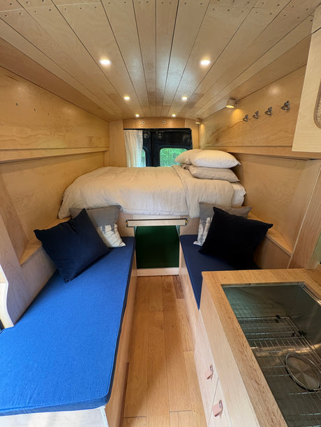 Step inside this cozy RV rental with natural wood walls, a comfy bed, blue cushions, and a functional kitchen sink.