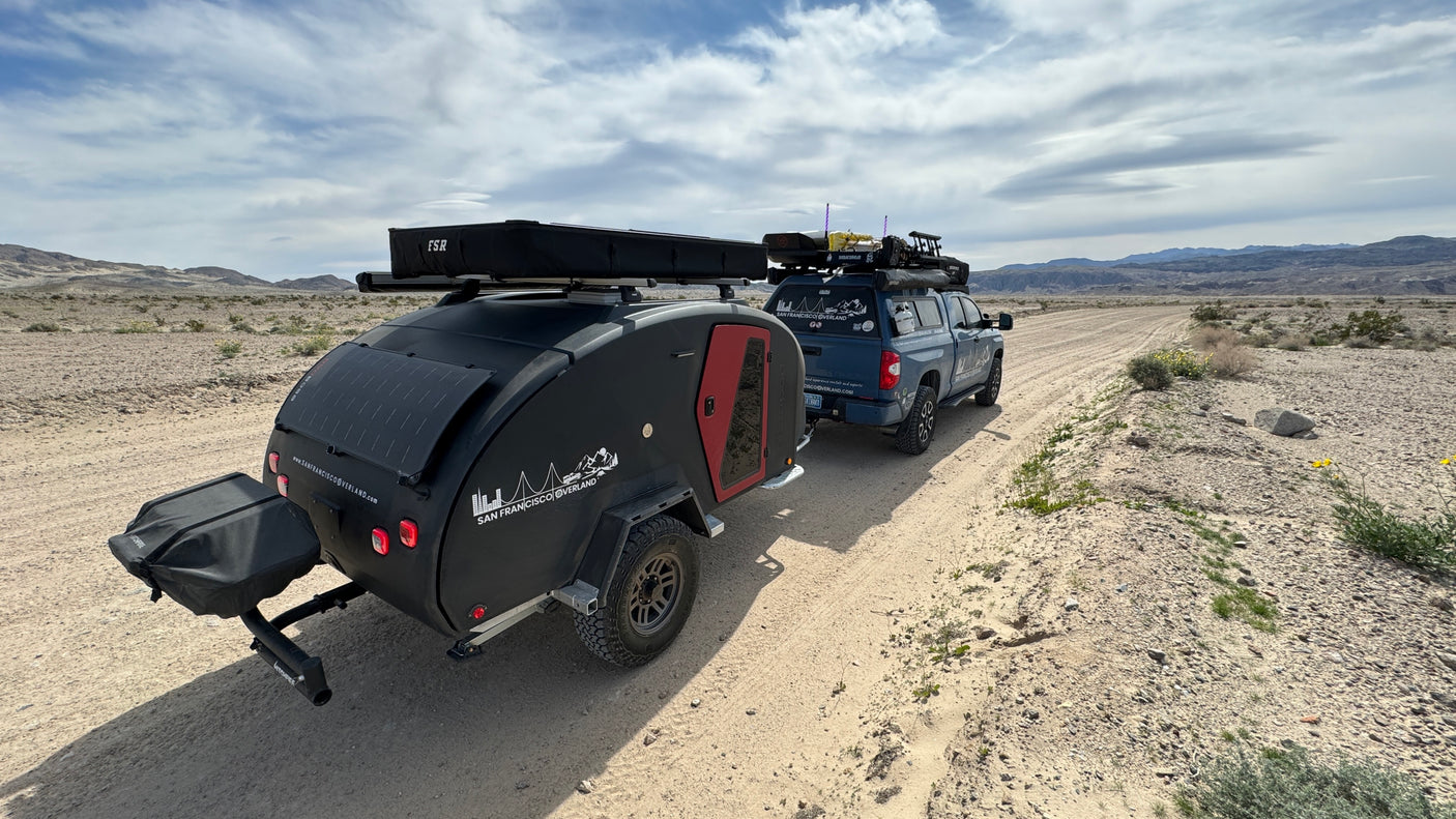This RV rental features a black overland teardrop trailer towed by a truck on a desert dirt road, under a cloudy sky.