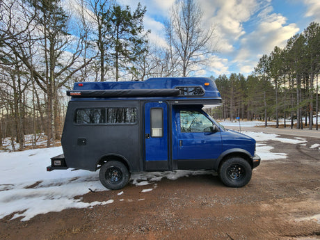 This rugged blue overland camper van, excellent for rv rental excursions, is parked on a snowy dirt road amidst winter trees.