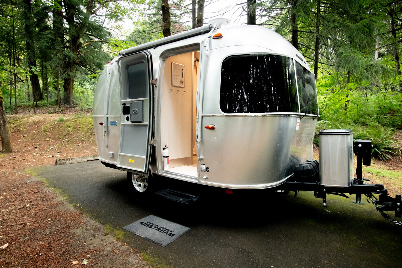A silver Airstream travel trailer RV rental with an open door, set in a peaceful, lush forest campsite.