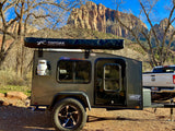 Teardrop camper RV rental with a Topoak awning, parked in a scenic desert landscape with towering canyon walls & bare trees.
