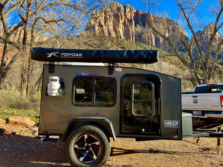 Teardrop camper RV rental with a Topoak awning, parked in a scenic desert landscape with towering canyon walls & bare trees.