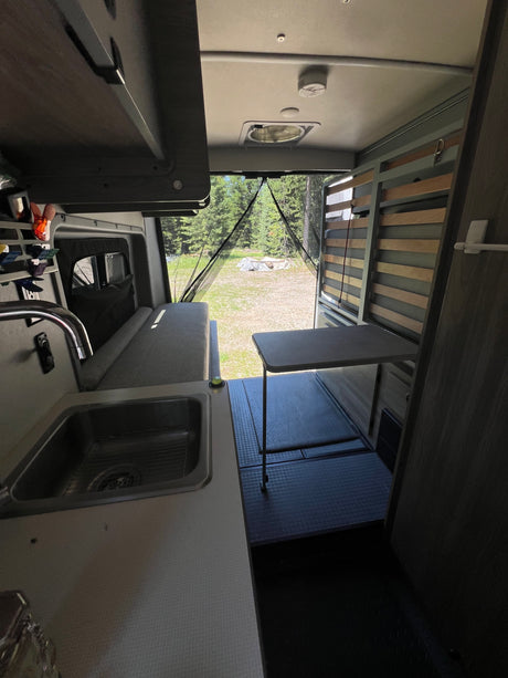 Spacious RV rental interior with kitchen sink, bed, movable table, and scenic outdoor view from open rear.