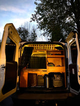 Cozy camper van interior seen from the rear, doors open. Custom wood build, warm string lights, & storage. Perfect RV rental.
