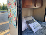 Inside an RV rental: a cozy bed with pillows, a vibrant curtain, and a view of the gravel campsite outside.
