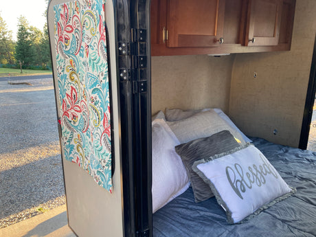 Inside an RV rental: a cozy bed with pillows, a vibrant curtain, and a view of the gravel campsite outside.