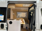 View inside an RV rental conversion in progress. Features new wood & white cabinetry, a roof vent, & driver's seat in a white camper van.