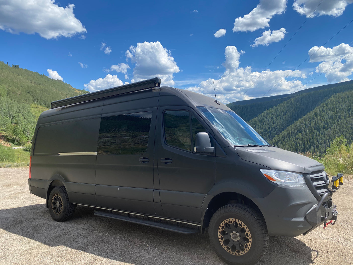 Black Mercedes Sprinter adventure van, an RV rental, parked on gravel. Features mountains, blue sky, and off-road tires.