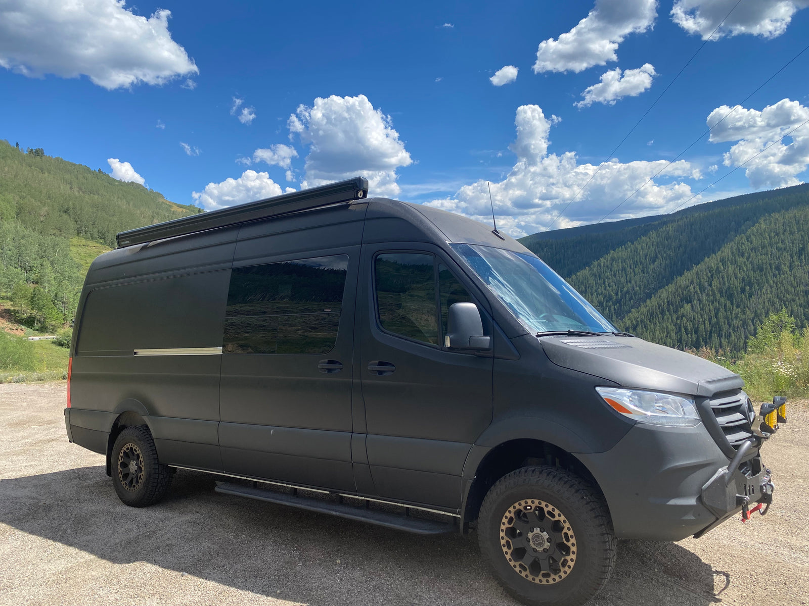 Black Mercedes Sprinter adventure van, an RV rental, parked on gravel. Features mountains, blue sky, and off-road tires.