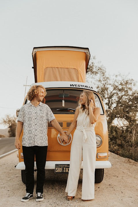 Happy couple by their RV rental, a vintage VW Westfalia camper van, holding hands & smiling. Perfect for adventure!