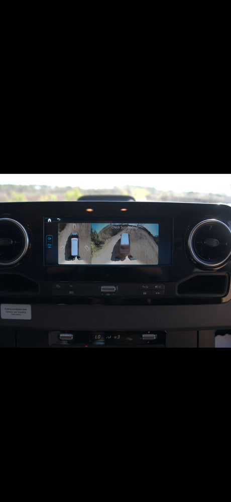 RV rental dashboard: infotainment screen shows 360-degree camera view ("Check Surroundings") of the vehicle on a dirt road.