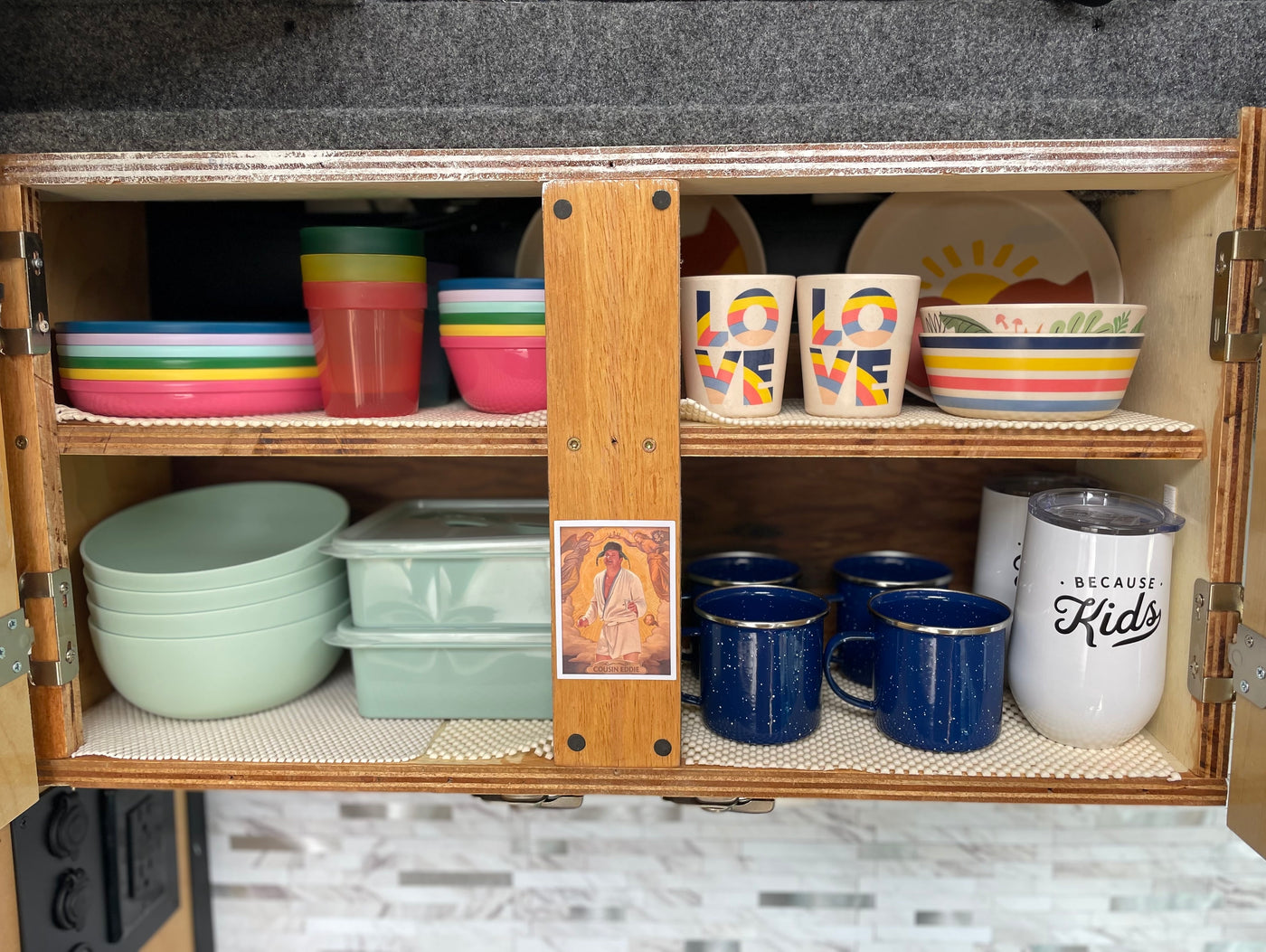 RV rental kitchen cabinet with colorful dishes & mugs, neatly organized for travel. Includes a Cousin Eddie pic. — photo 8