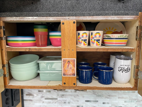 RV rental kitchen cabinet with colorful dishes & mugs, neatly organized for travel. Includes a Cousin Eddie pic.