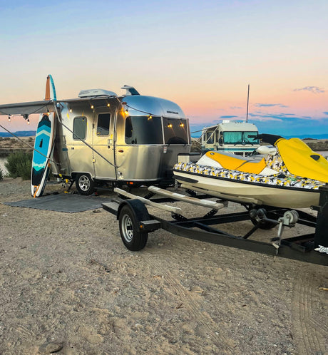 An Airstream RV rental, jet ski, and paddleboards are set up on sandy ground under a colorful sunset sky.
