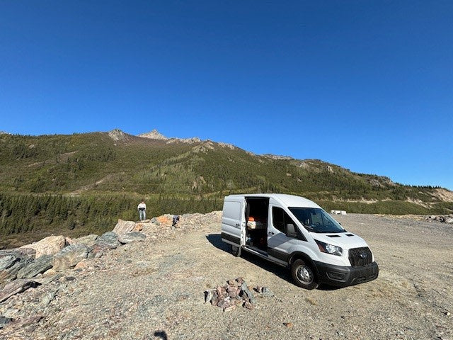 An RV rental parked on a mountain overlook with a person enjoying the scenic view of forested peaks under a blue sky.