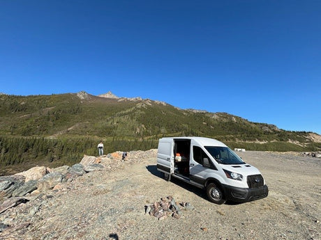 An RV rental parked on a mountain overlook with a person enjoying the scenic view of forested peaks under a blue sky.