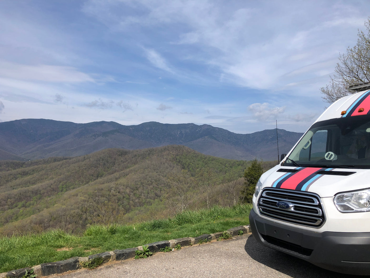 White Ford Transit RV rental van with Martini stripes at a mountain scenic overlook under a blue sky, showcasing the vast landscape.