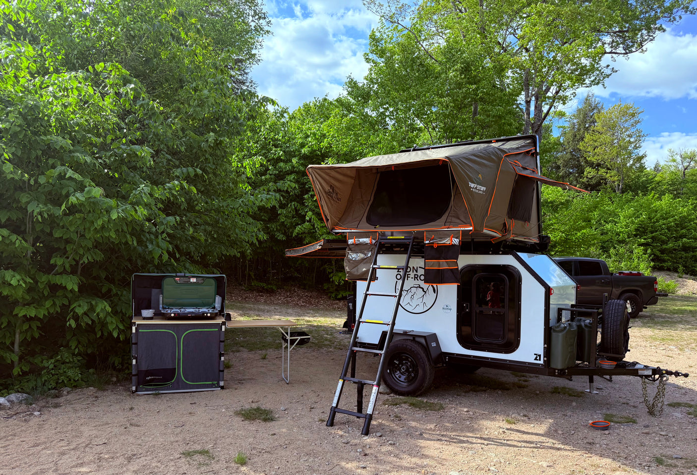 This RV rental teardrop trailer with rooftop tent and outdoor kitchen is set up for overland camping in a serene, green forest.