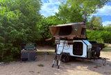 This RV rental teardrop trailer with rooftop tent and outdoor kitchen is set up for overland camping in a serene, green forest.
