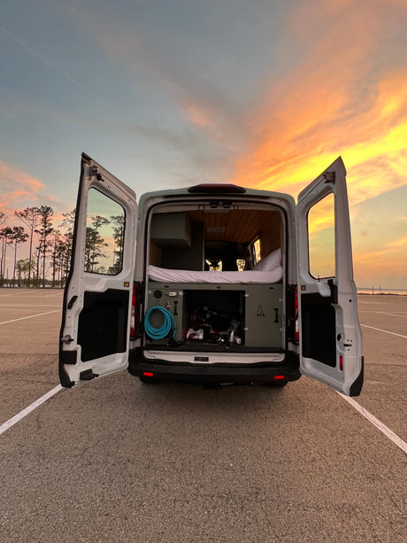 RV rental camper van's open rear reveals a cozy bed, interior storage, and gear. Golden sunset in the background.