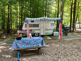 Charming RV rental camper under a festive awning with picnic tables in a serene woodland campsite.