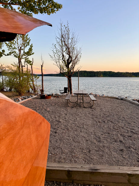 Peaceful lakeside RV rental spot with picnic table, fire pit, and beautiful lake view at sunset. Gravel ground, trees, and calm water.