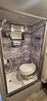 Compact RV rental bathroom with a combined shower and toilet, mirror, and storage shelves on tiled walls.