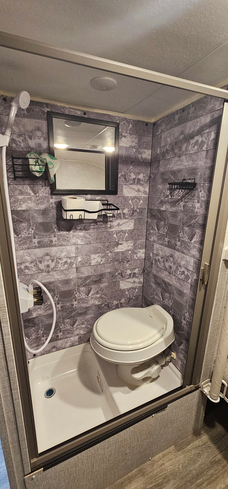 Compact RV rental bathroom with a combined shower and toilet, mirror, and storage shelves on tiled walls.