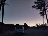 An RV rental van is silhouetted at dusk, nestled among tall pines against a colorful sky. Ideal for a peaceful nature escape.