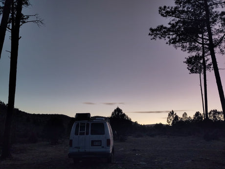 An RV rental van is silhouetted at dusk, nestled among tall pines against a colorful sky. Ideal for a peaceful nature escape.