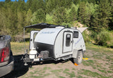 White Bushwhacker teardrop RV rental camper with awning extended in a scenic forest campground. Truck visible.