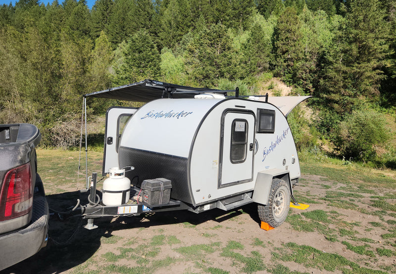 White Bushwhacker teardrop RV rental camper with awning extended in a scenic forest campground. Truck visible. — photo 7