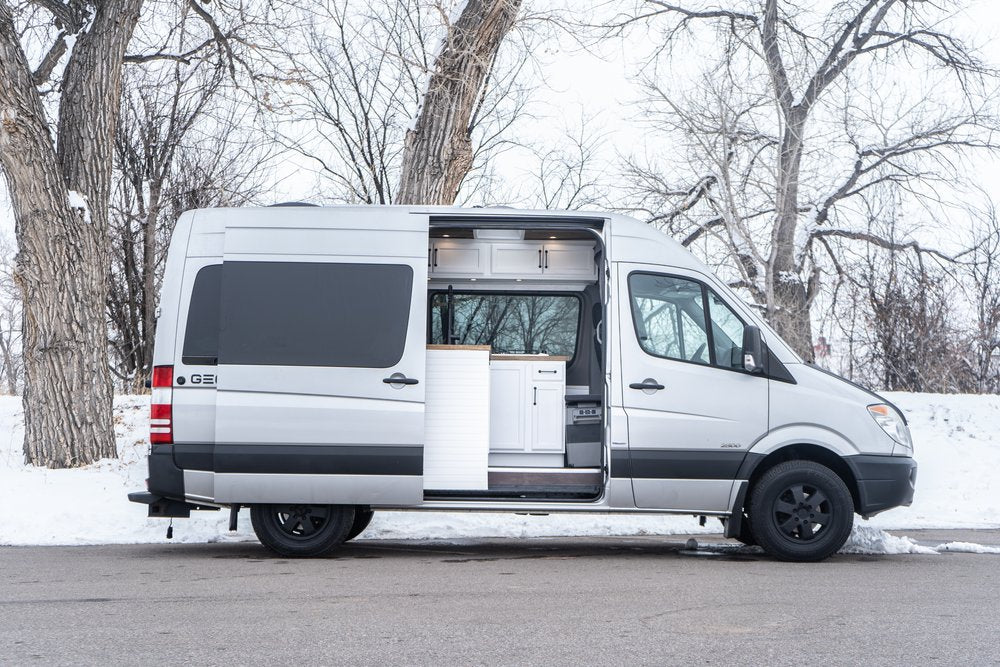 This silver Sprinter rv rental camper van is shown with its side door open, revealing a bright, modern white interior. Parked on a snowy day.