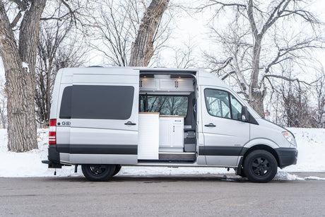 This silver Sprinter rv rental camper van is shown with its side door open, revealing a bright, modern white interior. Parked on a snowy day.