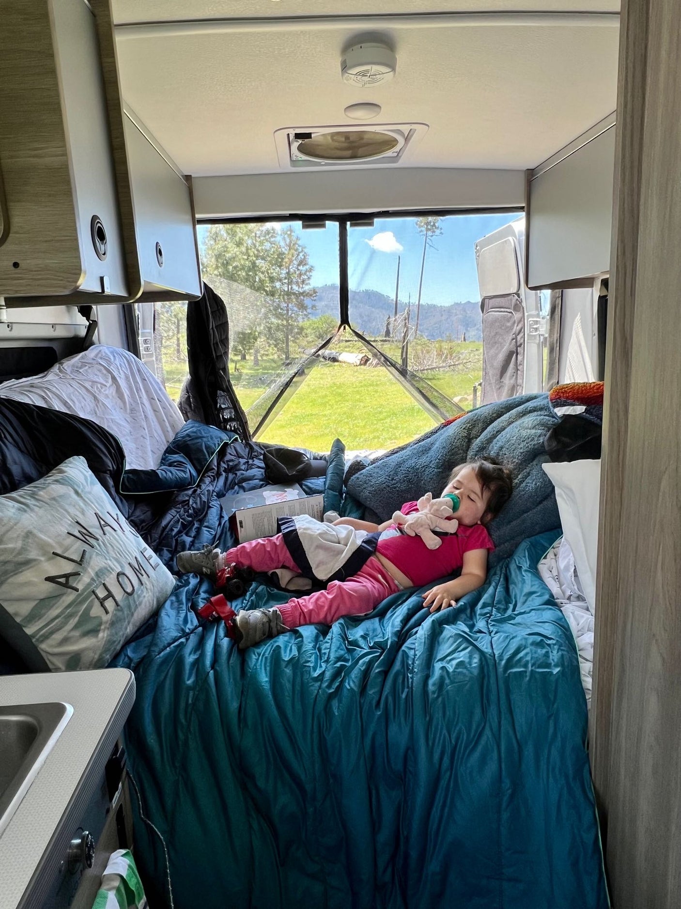 An RV rental interior with a child sleeping soundly, surrounded by a comfy bed, overlooking scenic nature views.