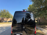 Black Mercedes-Benz Sprinter van, an Ultimate Coach perfect for rv rental, parked on a sunny street reflecting trees and houses.