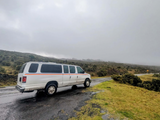 White RV rental van on a wet road, surrounded by misty hills and green scrubland. Perfect for a nature road trip.