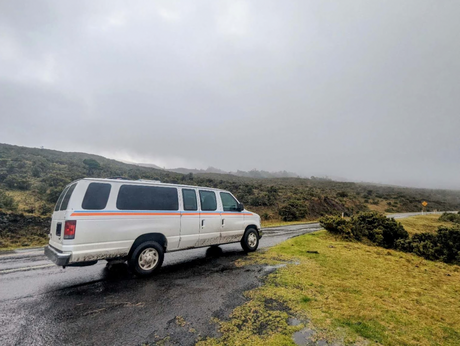 White RV rental van on a wet road, surrounded by misty hills and green scrubland. Perfect for a nature road trip.