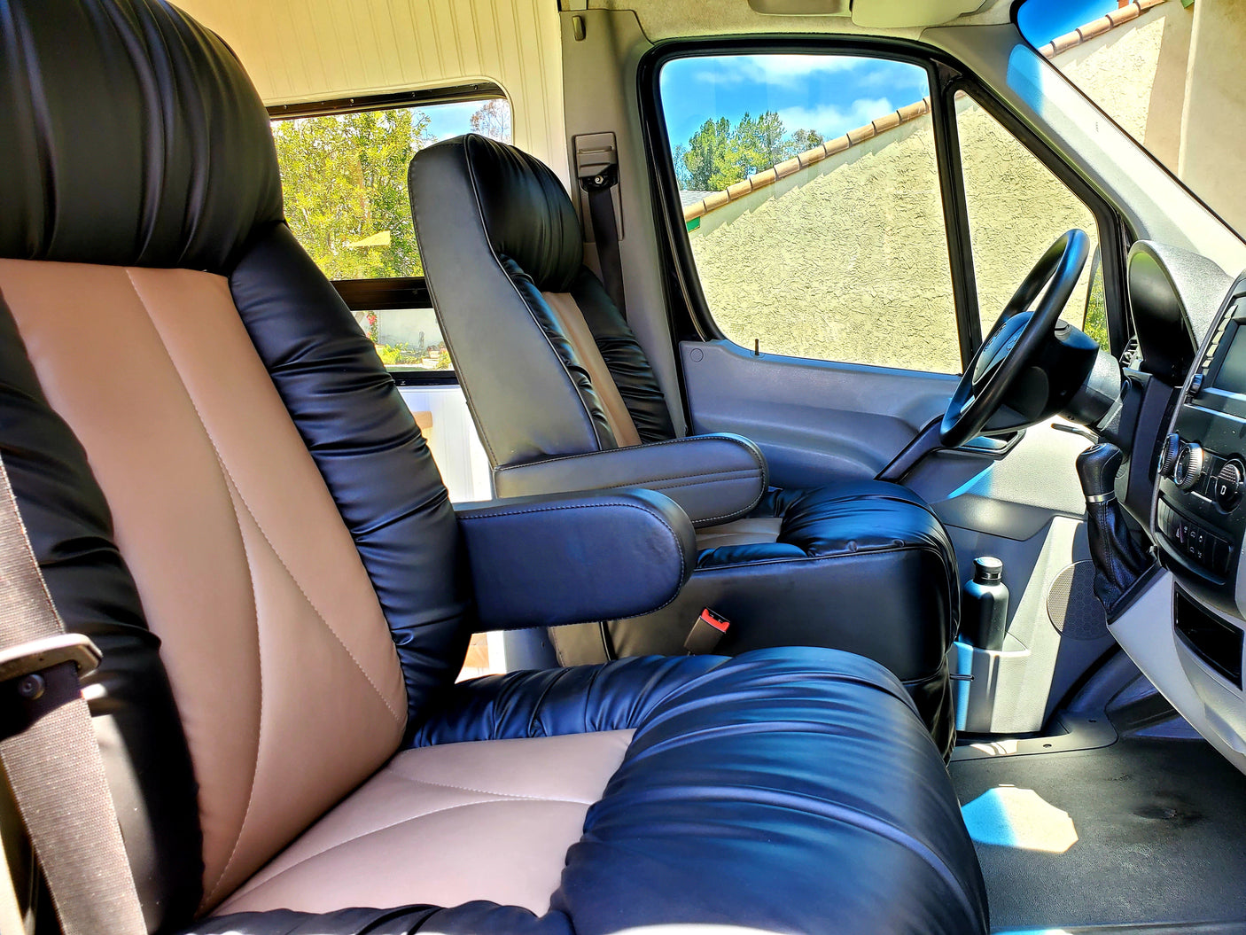 View inside an rv rental: plush black and tan front seats with armrests. Bright windows reveal a sunny outdoor scene.
