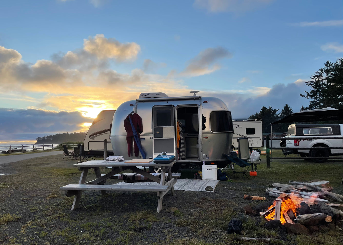An RV rental Airstream at a coastal campsite with a crackling campfire, sunset clouds, and ocean view. Ideal for travel.