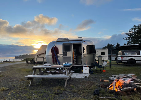 An RV rental Airstream at a coastal campsite with a crackling campfire, sunset clouds, and ocean view. Ideal for travel.