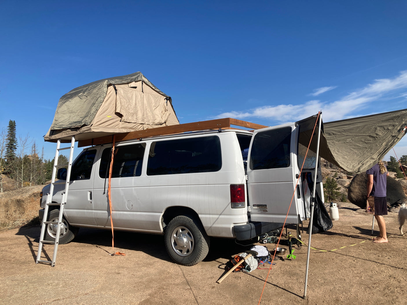 RV rental ready for adventure: A white van with a rooftop tent and awning set up at a sunny outdoor campsite with a person & dog.
